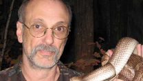 Man wearing glasses and a camouflage shirt holding a large snake wrapped around his arm.
