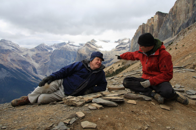 Two people wearing outdoor clothing sit and point at rocks on a mountainous trail with snow-capped peaks in the background.