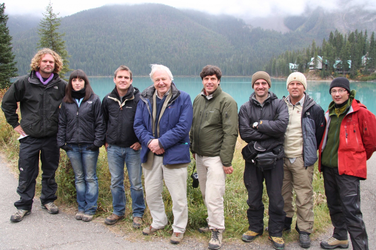 A group of eight people standing outdoors on gravel with a lake and misty forested hills in the background.