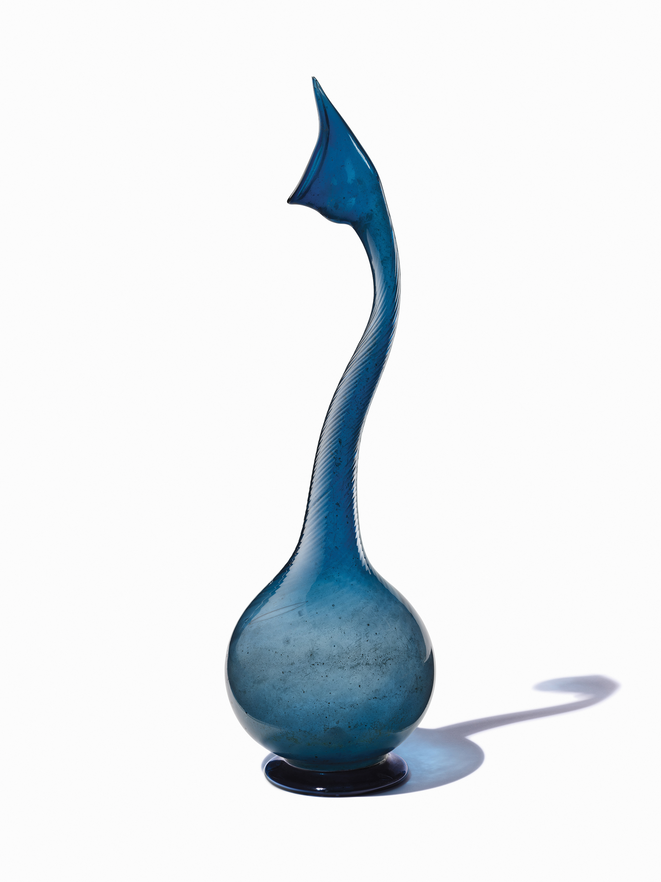 Tall, slender blue glass vase with a curved neck and bulbous base casting a shadow.