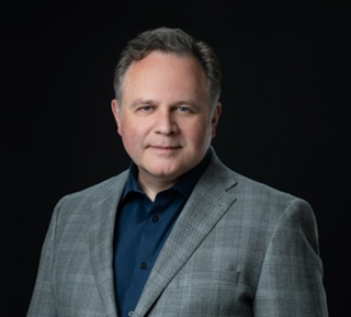 Portrait of a man with short gray hair wearing a gray checked blazer and a dark blue shirt against a black background.