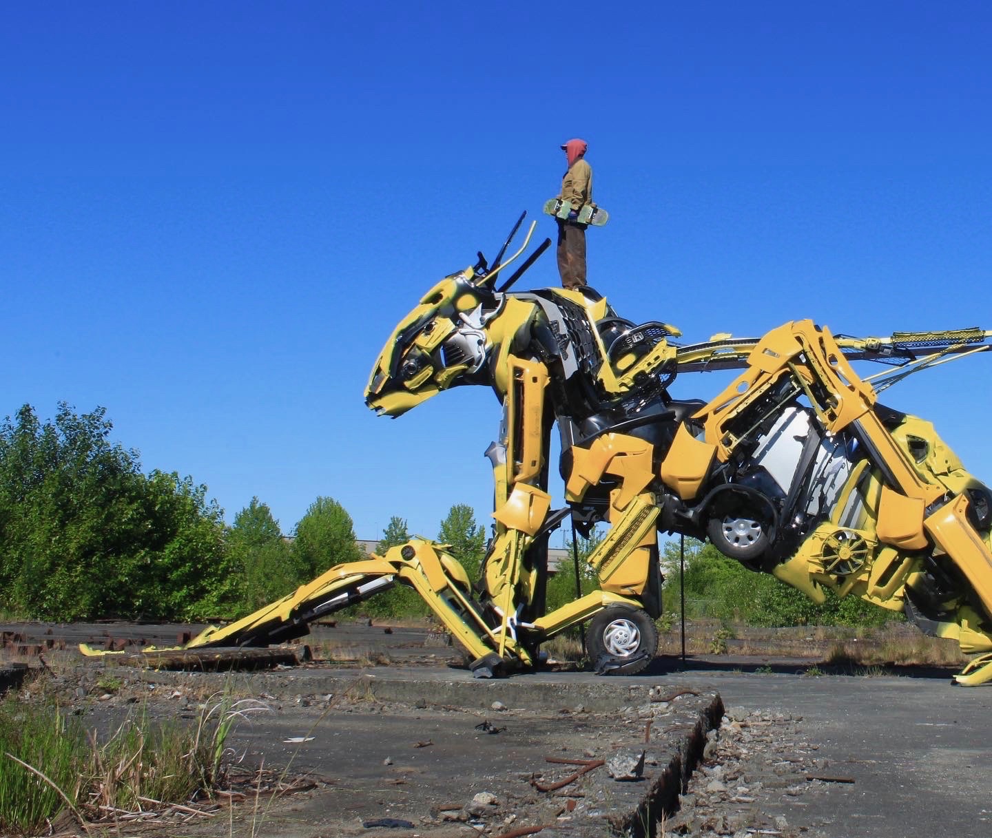 Grande sculpture de dinosaure mécanique jaune fabriquée à partir de pièces industrielles sur une plate-forme en bois, en plein air, sous un ciel bleu clair.