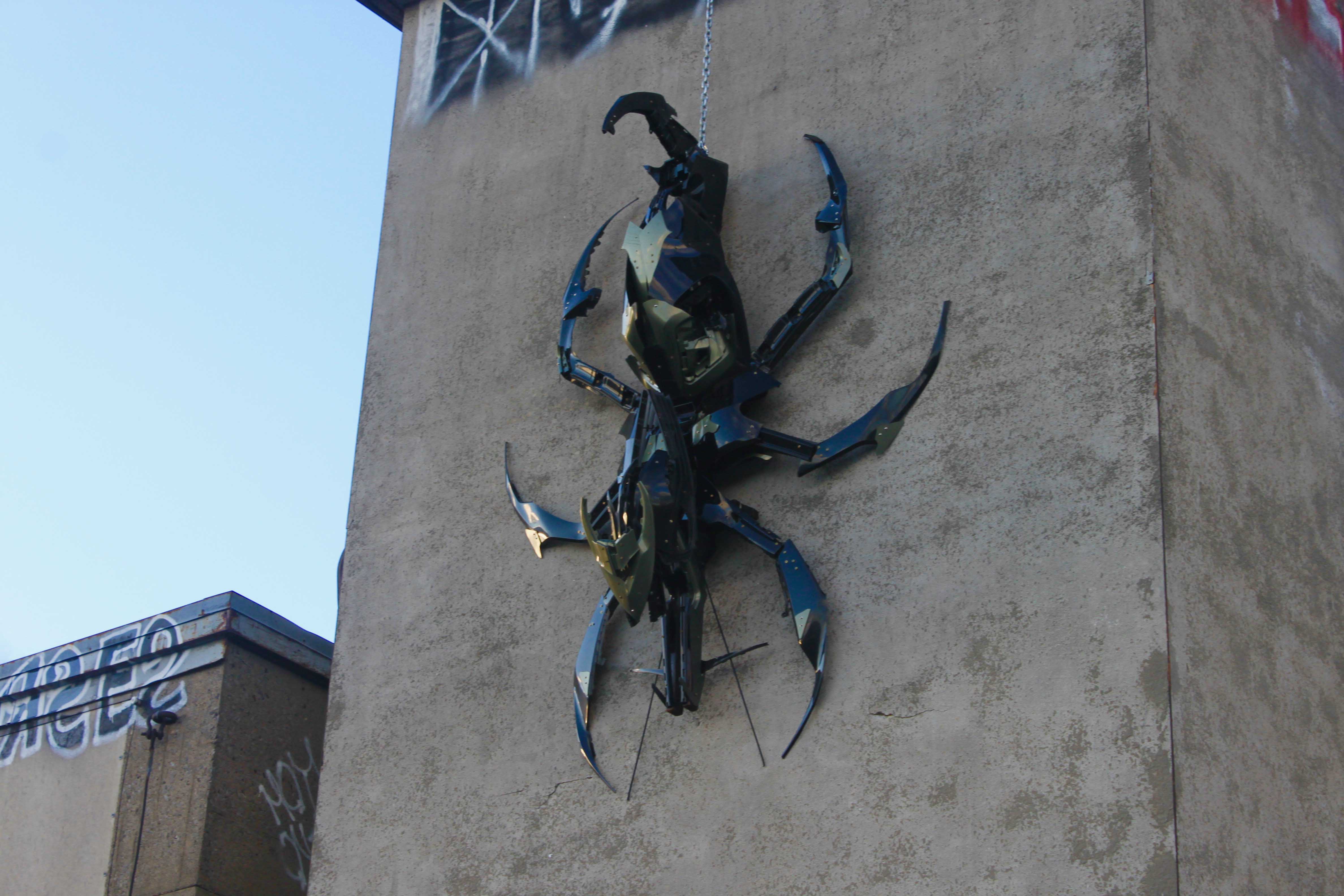 Grande sculpture de scarabée en métal bleu et noir montée sur le mur d'un bâtiment en béton beige.