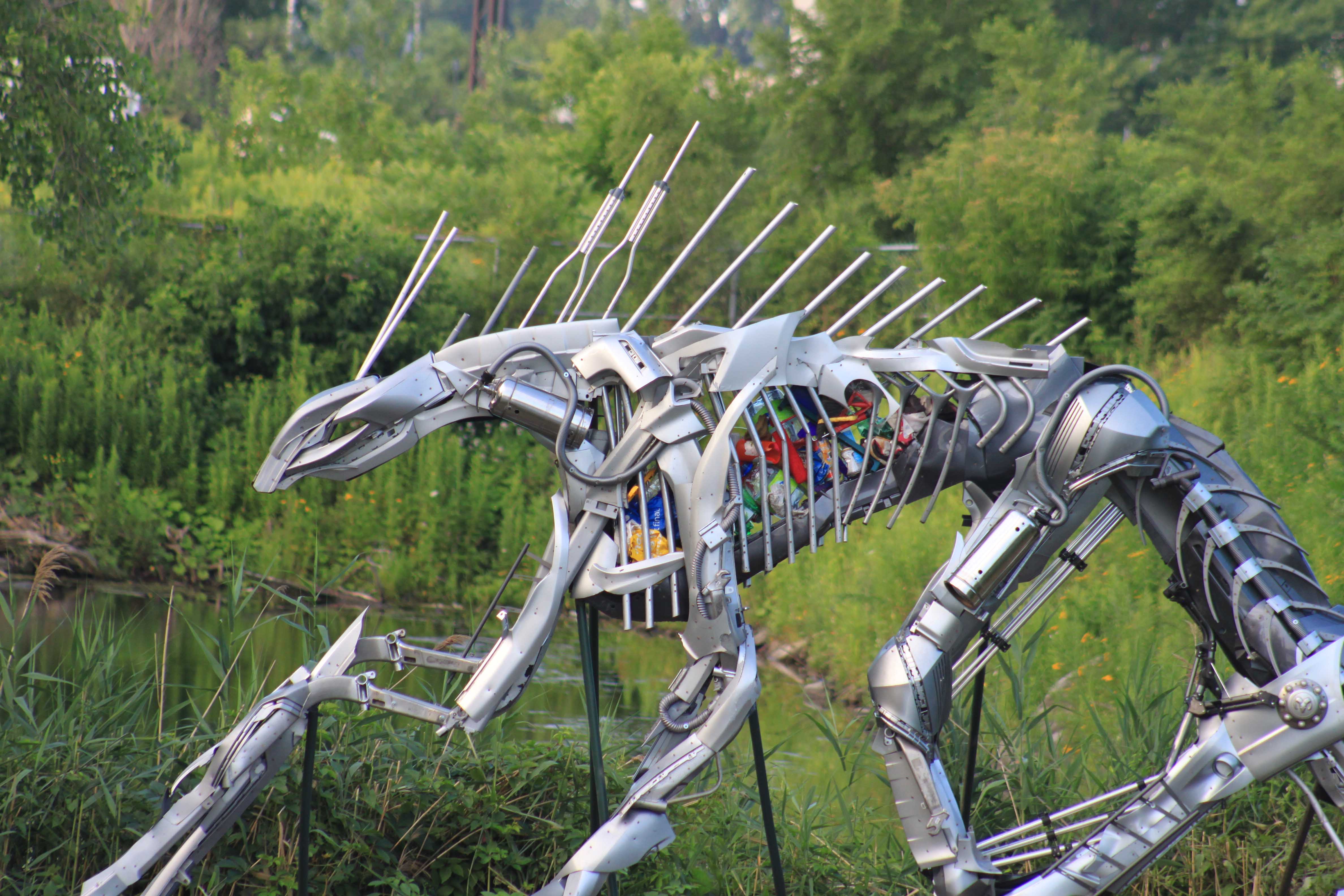 Grande sculpture métallique d'un insecte aux multiples pattes et antennes, placée en extérieur avec de la verdure en arrière-plan.