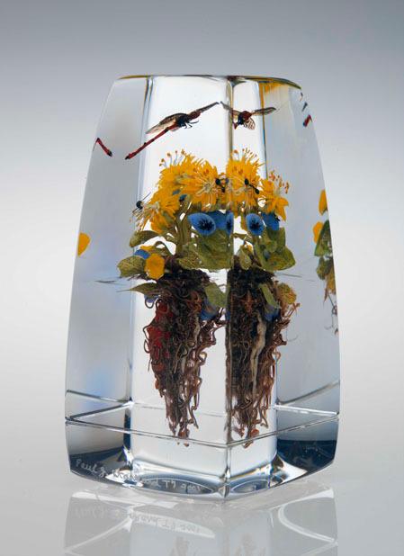 Yellow flowers with green leaves and brown roots encased in a clear rectangular resin block.