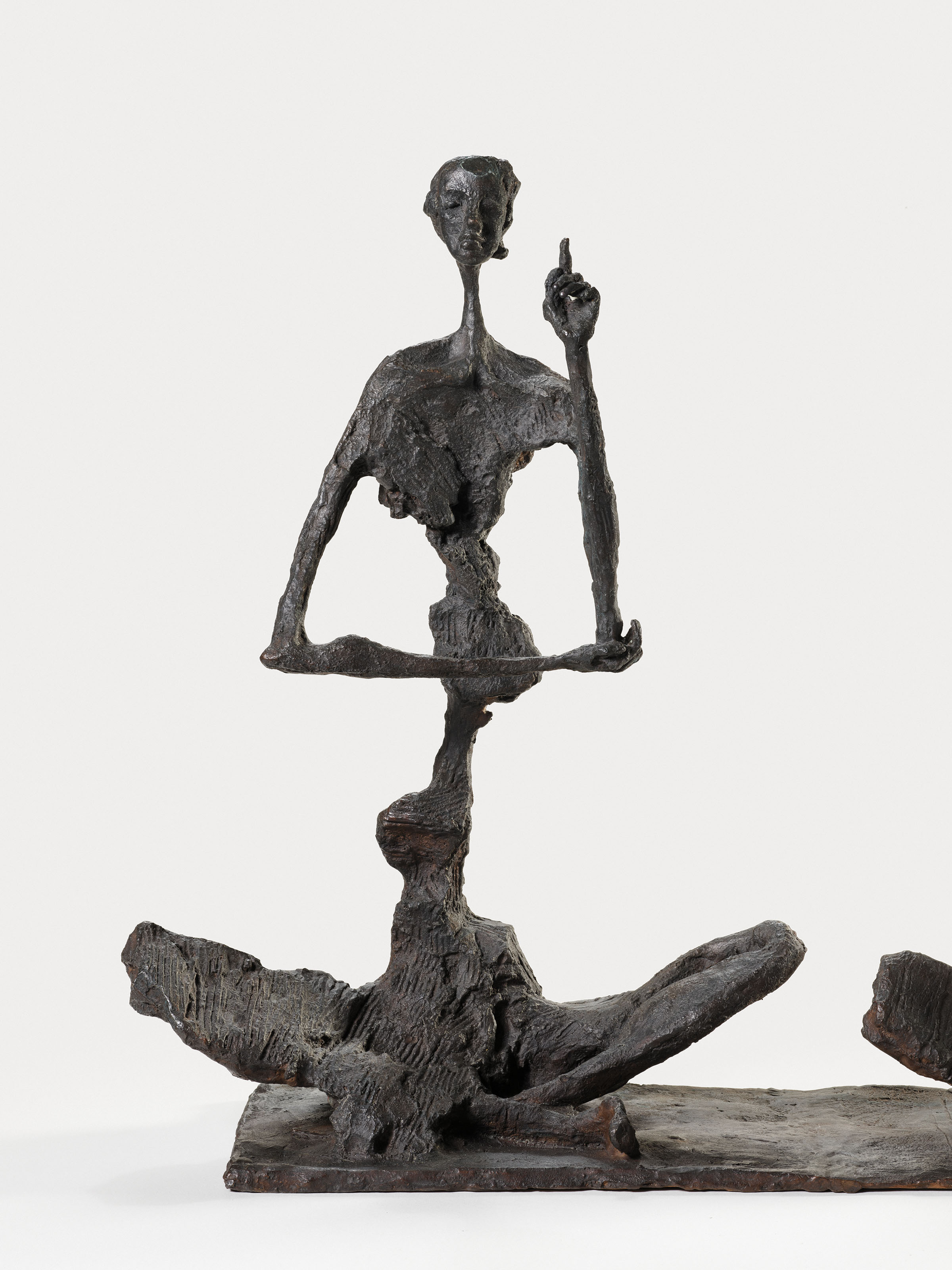 Bronze sculpture of two elongated, abstract figures stacked vertically, with the upper figure seated cross-legged and raising one hand.