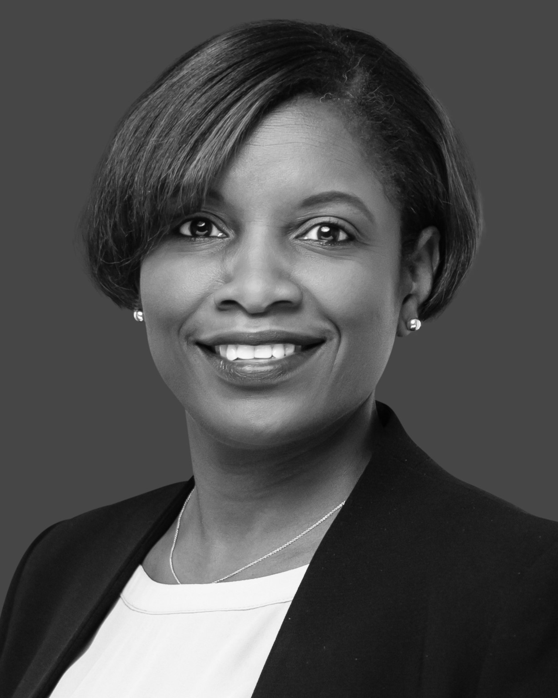 Portrait of a smiling woman with short hair wearing a dark blazer and white top, against a gray background.