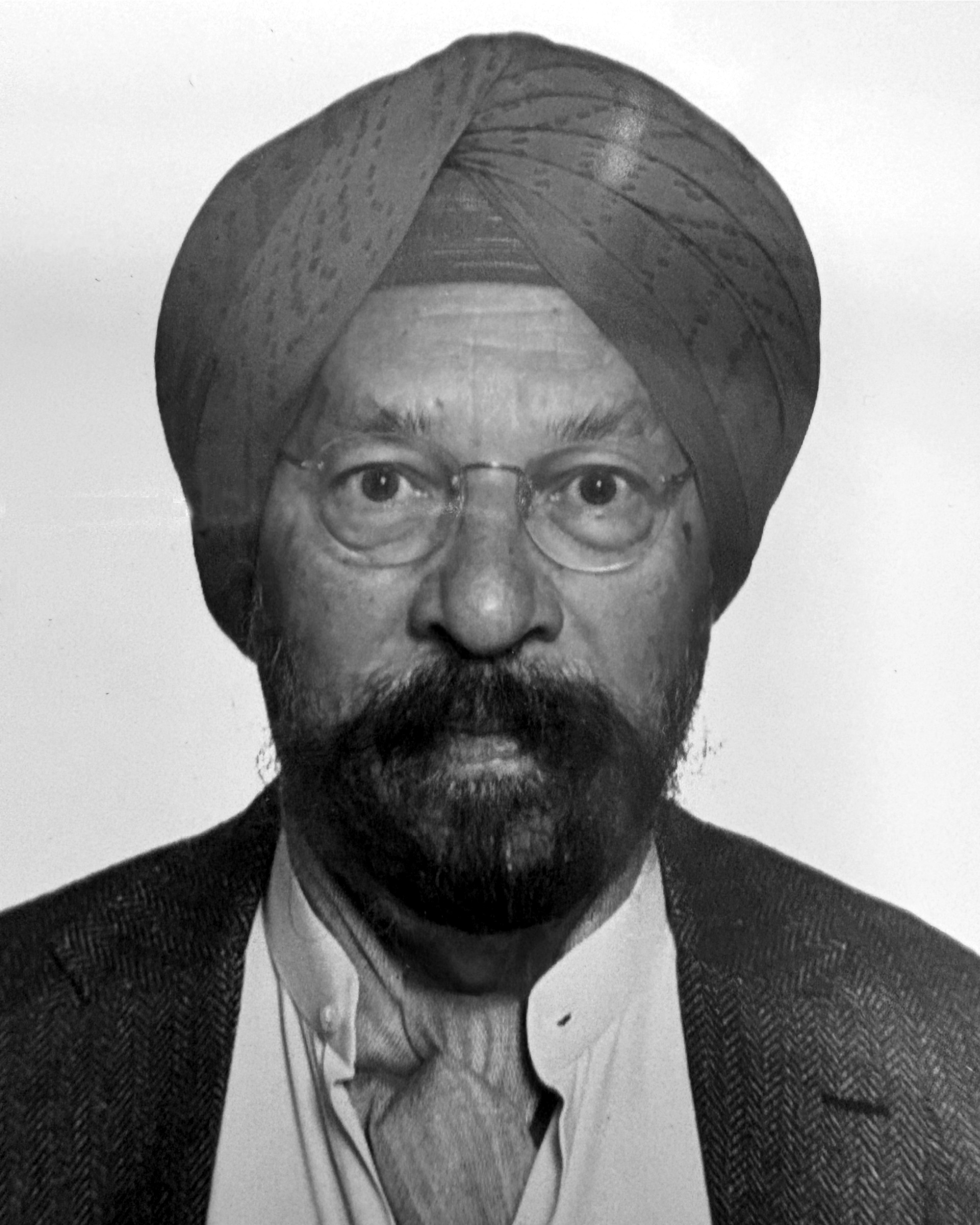 Portrait of an older man wearing glasses, a turban, and a dark blazer over a light shirt.
