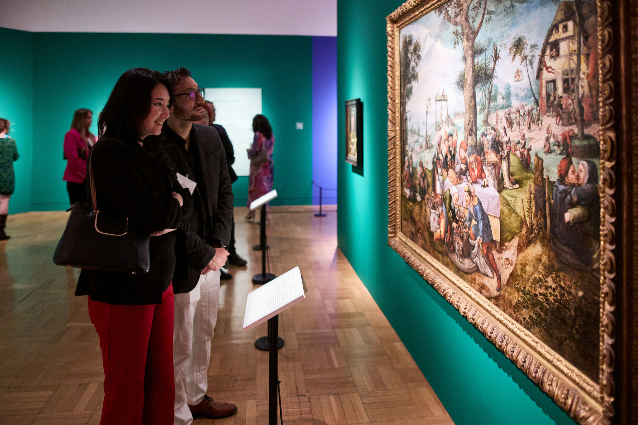 Two visitors closely observing a large framed painting displayed on a teal gallery wall.