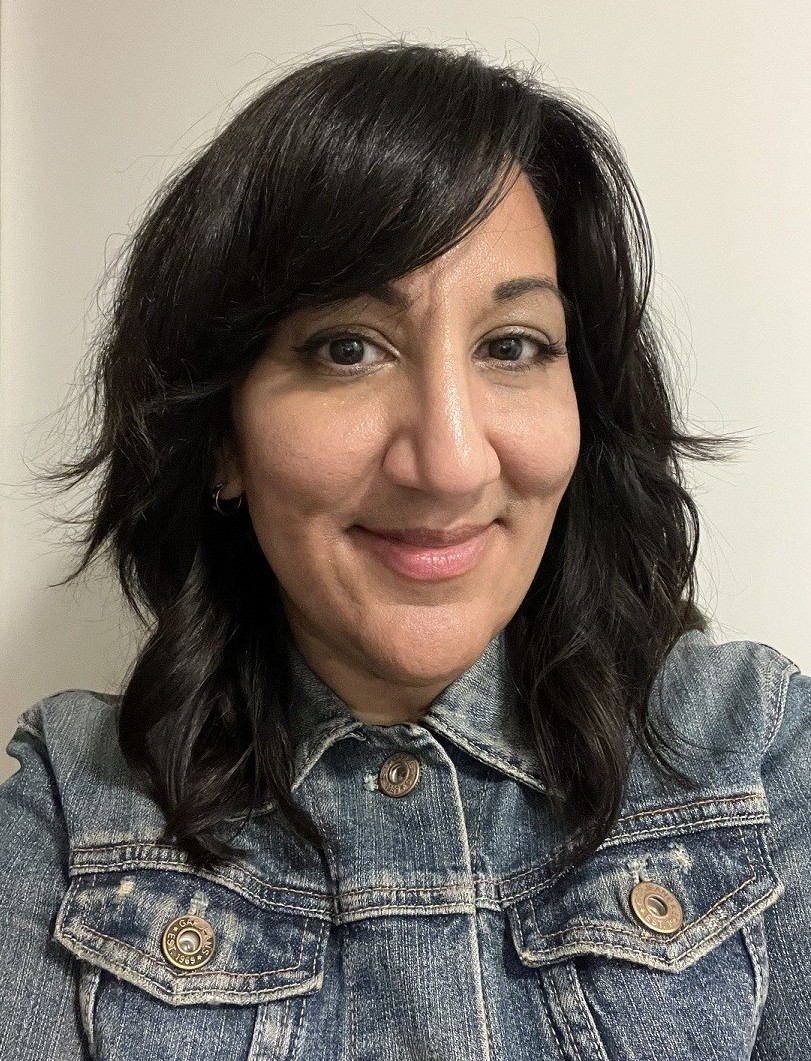 Woman with shoulder-length dark hair wearing a blue denim jacket smiling against a plain light background.