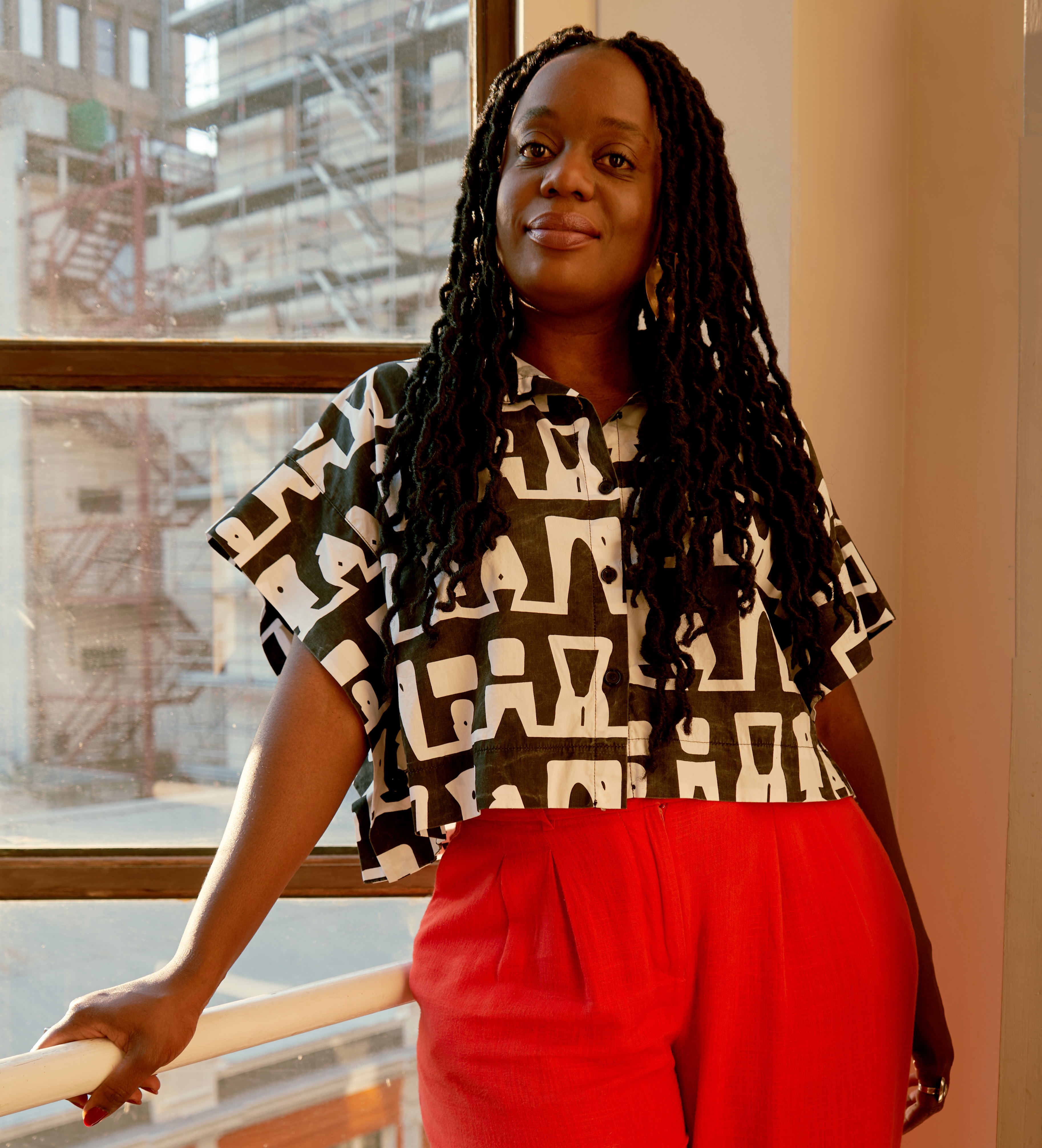 Woman with long braided hair wearing a black-and-white patterned top and red pants standing by a window with city buildings visible outside.