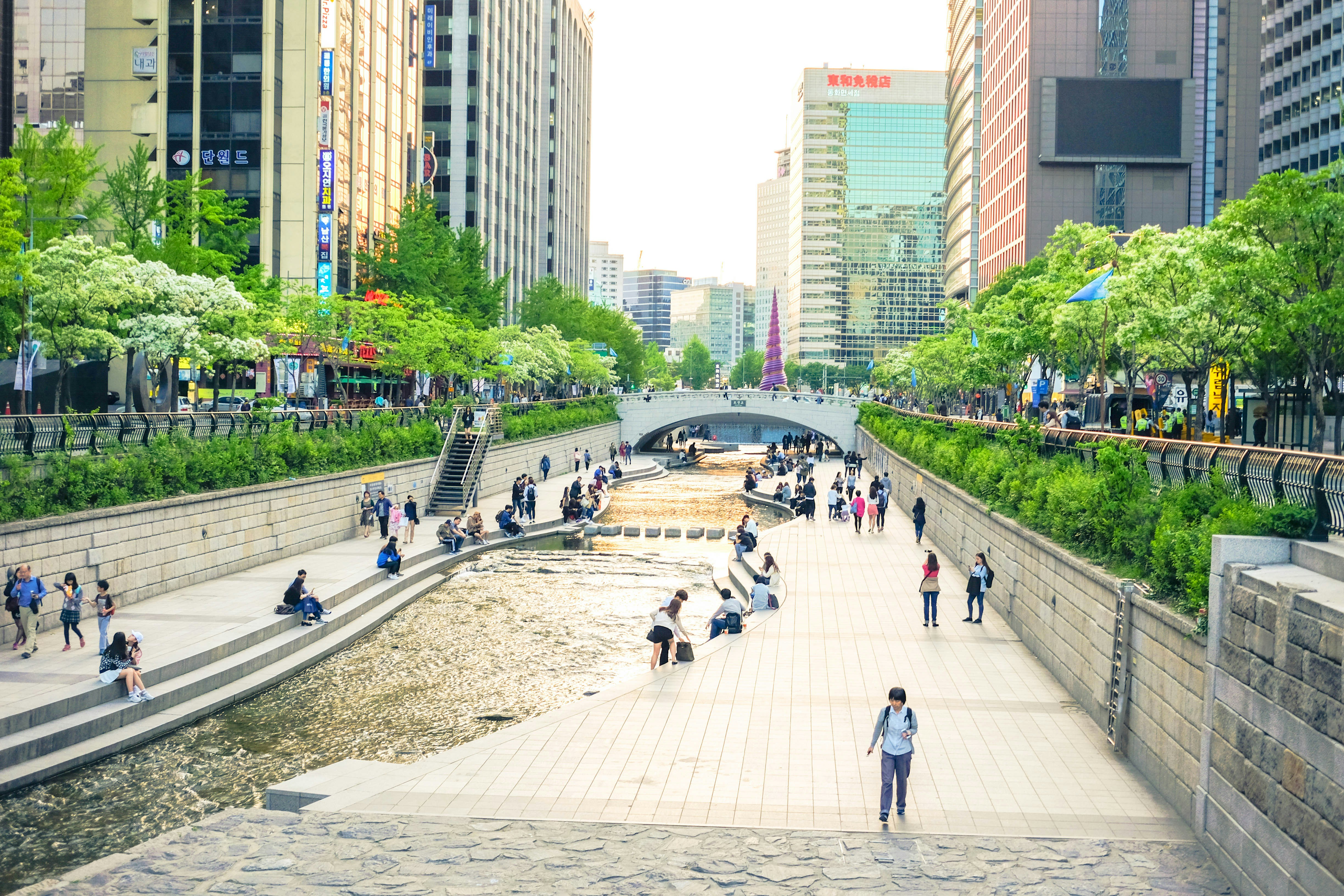 Urban waterway with stone steps on both sides, people walking and sitting along the stream in a city setting.