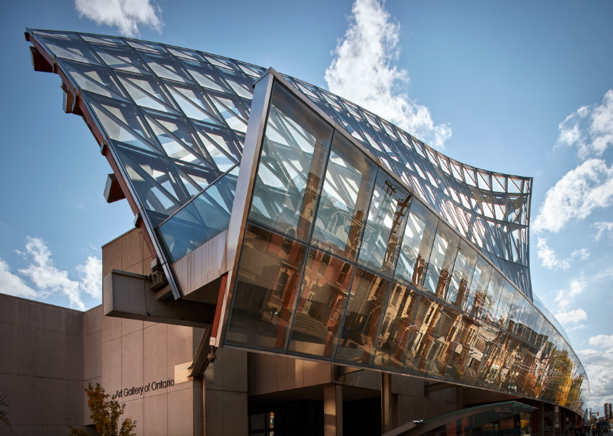 Exterior of the Art Gallery of Ontario featuring a striking curved glass façade with steel framing, reflecting nearby buildings under a blue sky.