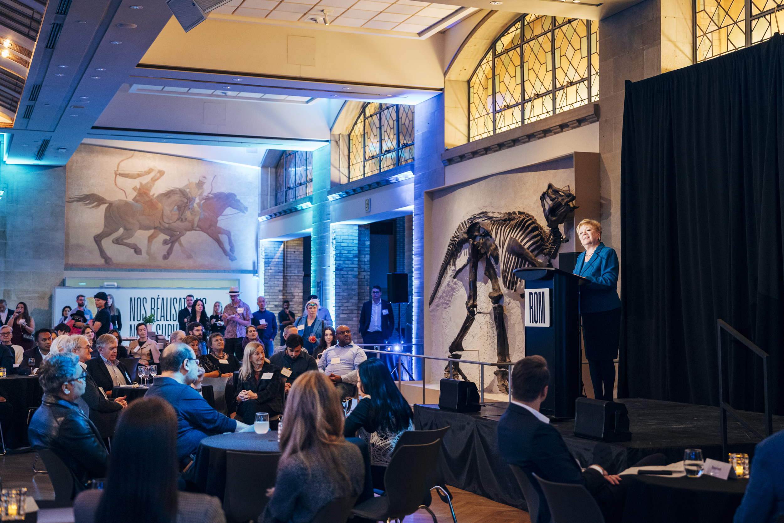 Audience seated in a large room listening to a speaker addressing the crowd from a stage with a large dinosaur skeleton.