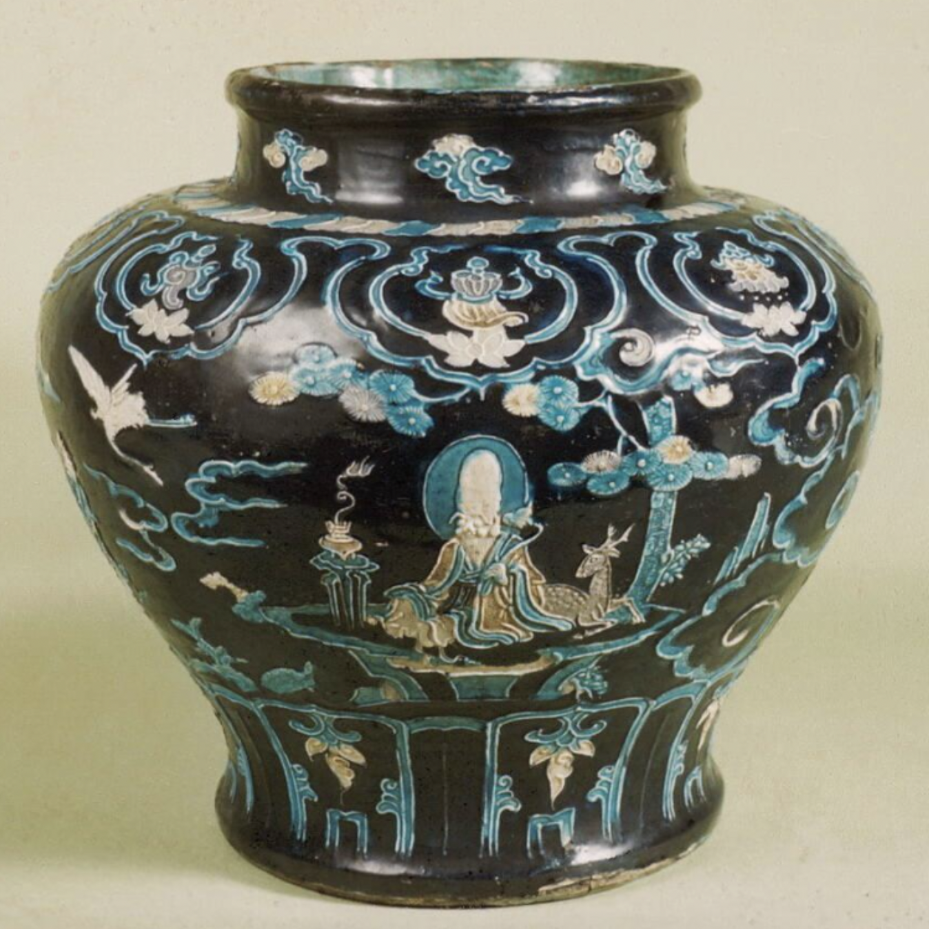 Decorative ceramic jar with a dark, glossy glaze and rounded body. The surface is richly ornamented with raised, multicolored designs in turquoise, blue, white, and pale accents, depicting robed human figures seated or standing in a stylized landscape with clouds, waves, floral motifs, and small animals. Curving bands and framed panels divide the scenes around the vessel, with additional decorative patterns encircling the neck and base. The jar is photographed against a light, neutral background.