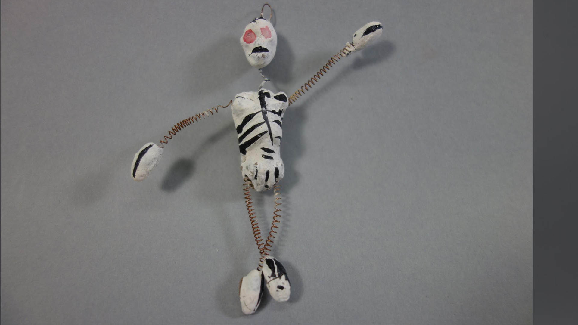 Petite figurine artisanale au corps blanc peint de marques noires, à la bouche rouge et aux yeux orange, posée sur un fond gris.