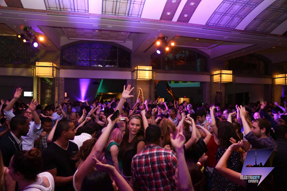 Crowd of people dancing with raised hands under decorative ceiling and multicolored stage lights.