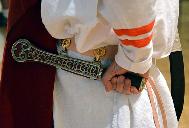 Close-up of a person wearing a white garment with orange stripes, holding a decorative sheathed dagger behind their back.