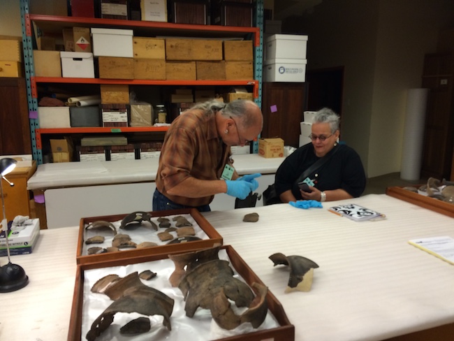 Two men examine large fossilized bones on a table in a room with shelves holding boxes in the background.