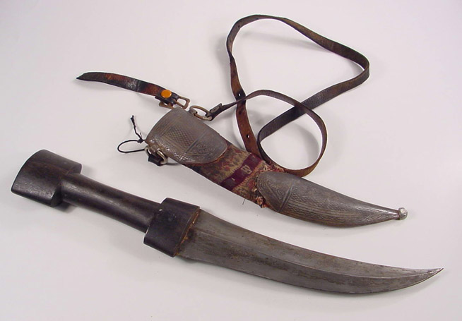 Curved knife with dark handle and sheath decorated with fabric and attached leather strap.