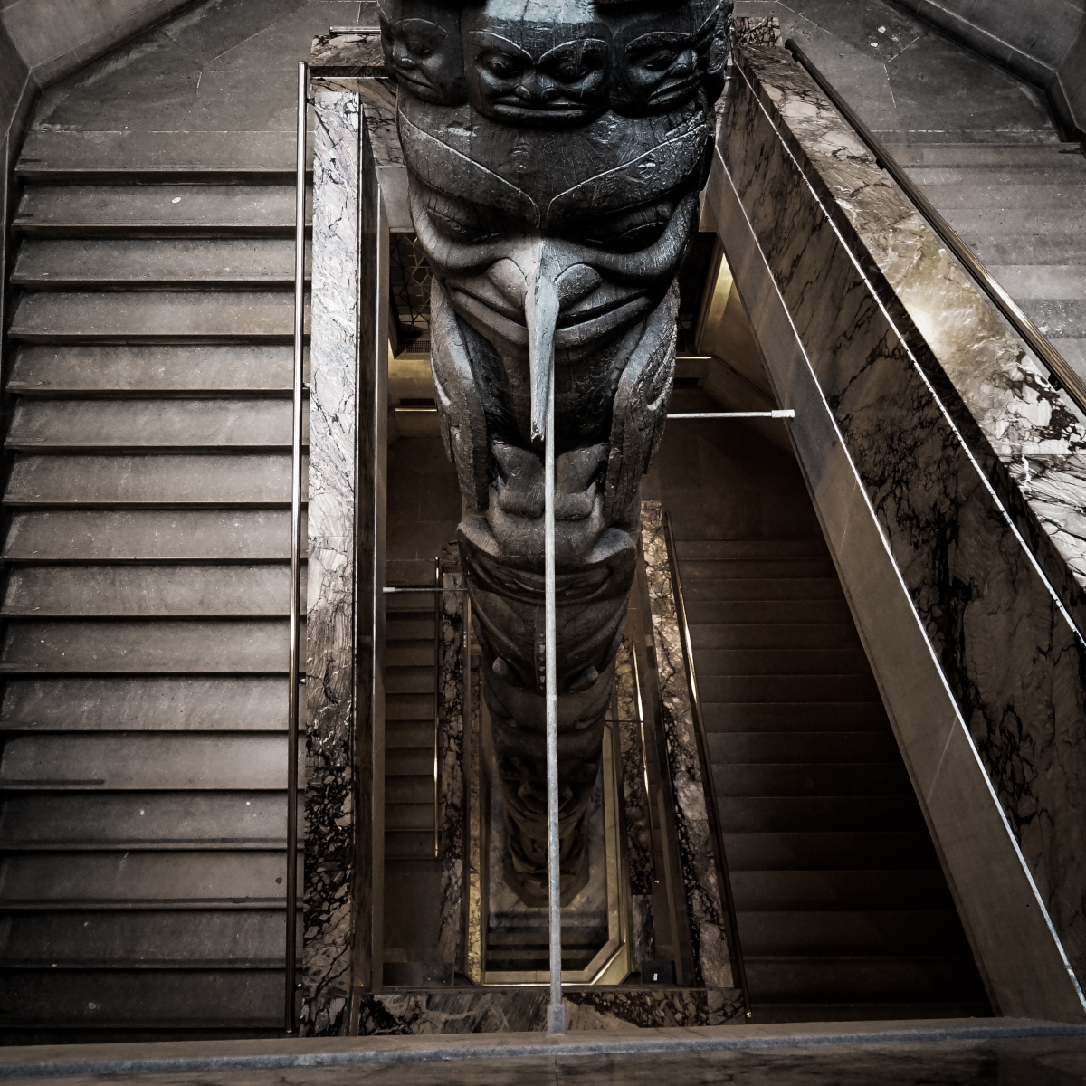 Top-down view of a carved totem pole placed in the middle of a stairwell with gray concrete stairs on either side.