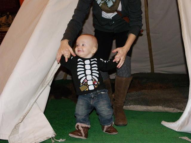 A baby wearing a black outfit with a white skeleton print and brown boots is standing on green flooring, supported by an adult.