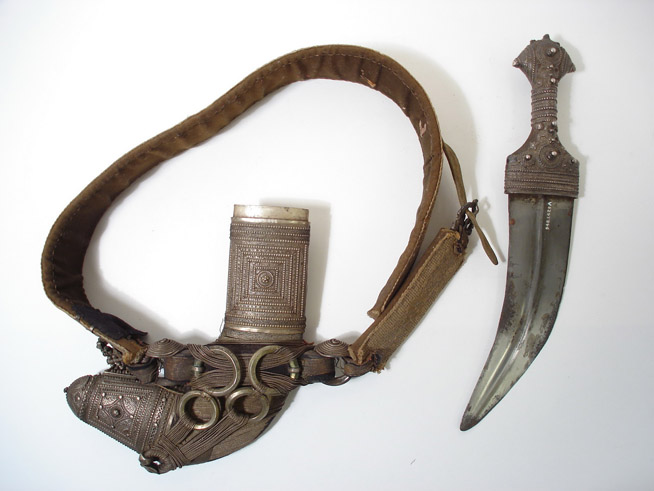 A curved dagger with an ornate handle and a leather sheath attached to a wide leather belt with metal cylindrical decorations.