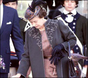 A woman in a dark coat with embroidery and matching hat holds a black handbag and wears black gloves.