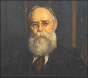 Portrait of an older man with glasses, a bald head, and a full white beard, dressed in a dark suit.