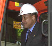Man in a suit and tie wearing a white hard hat with a red logo and standing inside construction machinery.