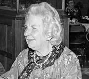 Elderly woman with curly white hair, wearing a patterned scarf and floral blouse, sitting and smiling, black and white photo.