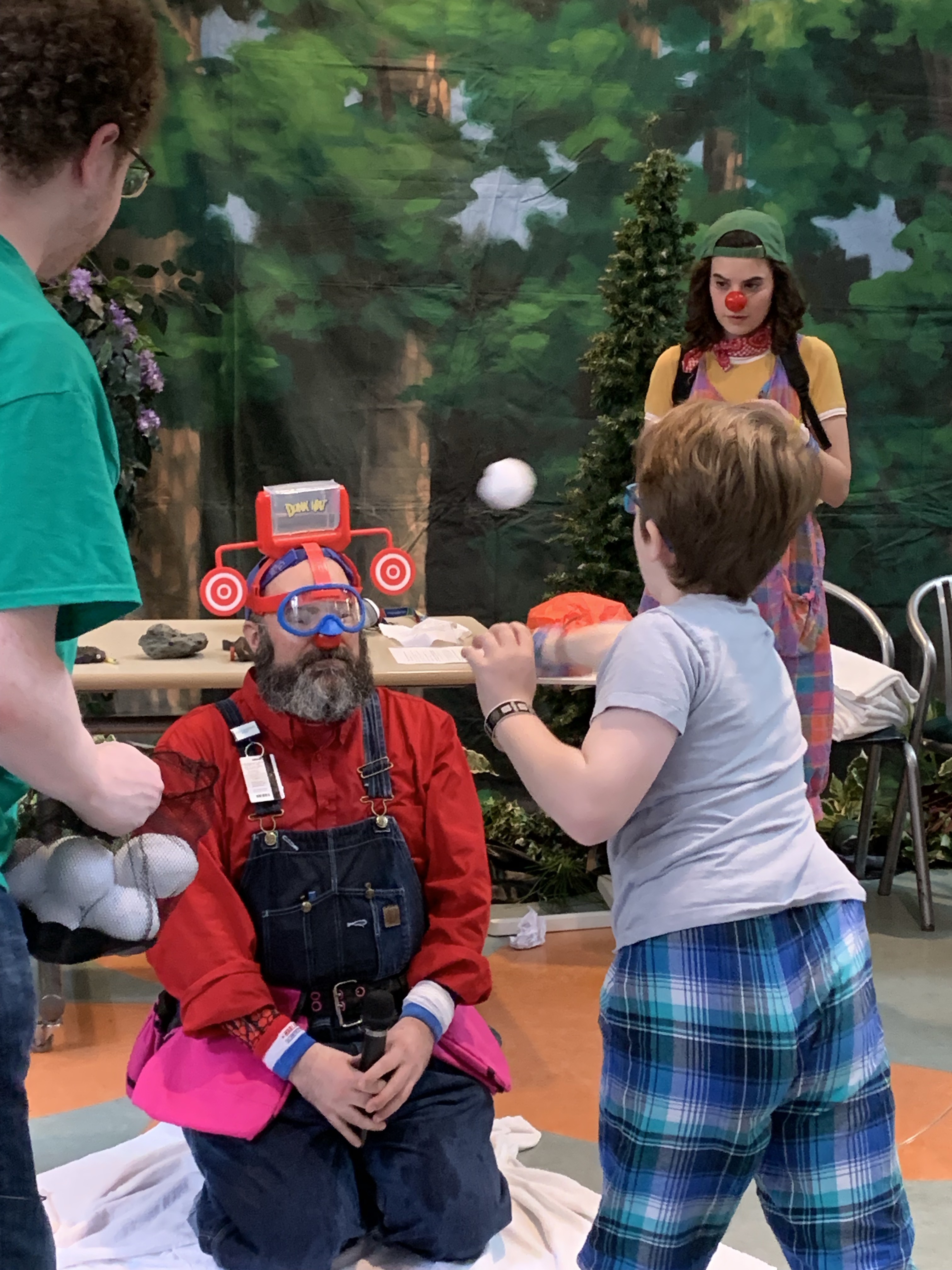 Three children playing outdoors on a forest-themed play mat; one child in a red shirt and goggles holds a toy bat, another child in blue pants swings a bat, and a third child in green watches.