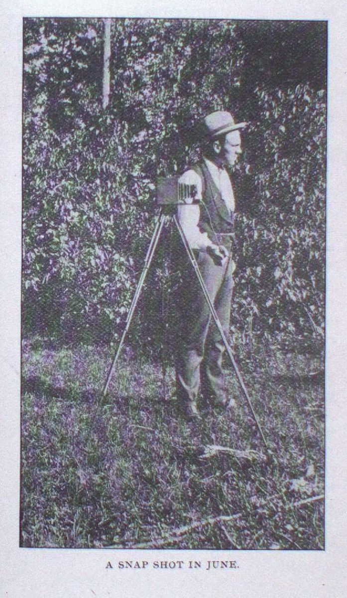 A man wearing a hat holds a long camera tripod, standing in front of dense foliage.