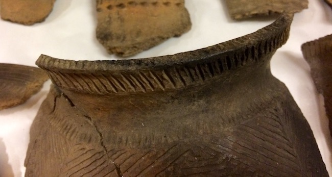 Close-up of a brown pottery vessel with a flared rim and incised linear and triangular patterns.