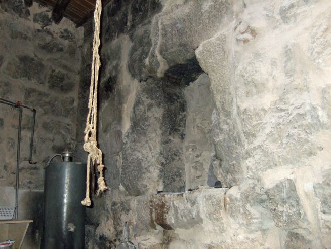 A rough stone wall with a thick rope hanging down in front of it and a large dark cylindrical object nearby.
