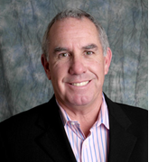Portrait of a smiling middle-aged man with short gray hair, wearing a black blazer and a white shirt with red stripes.