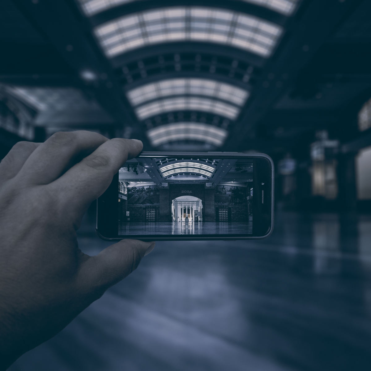 A hand holds a smartphone capturing the interior of a large hall with a high vaulted glass ceiling and symmetrically arranged columns.