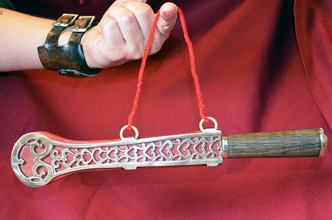 Hand holding a decorated metal sword guard with red string and a dark handle against a red fabric background.