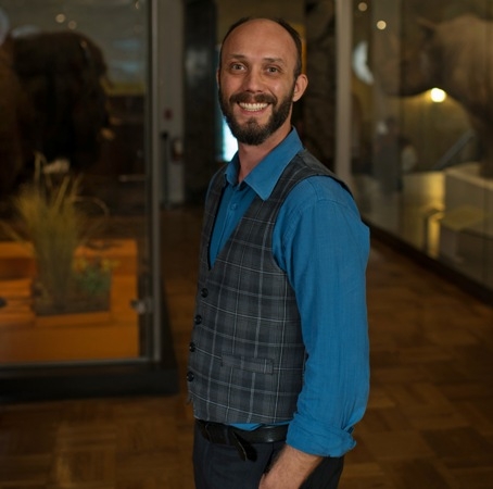 Portrait of a smiling man with a beard wearing a blue shirt and gray plaid vest in a dimly lit indoor setting.