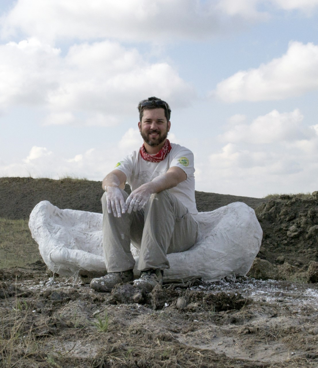 A man in light-colored clothes sits on the ground in front of a large, irregular white stone or fossil outdoors.