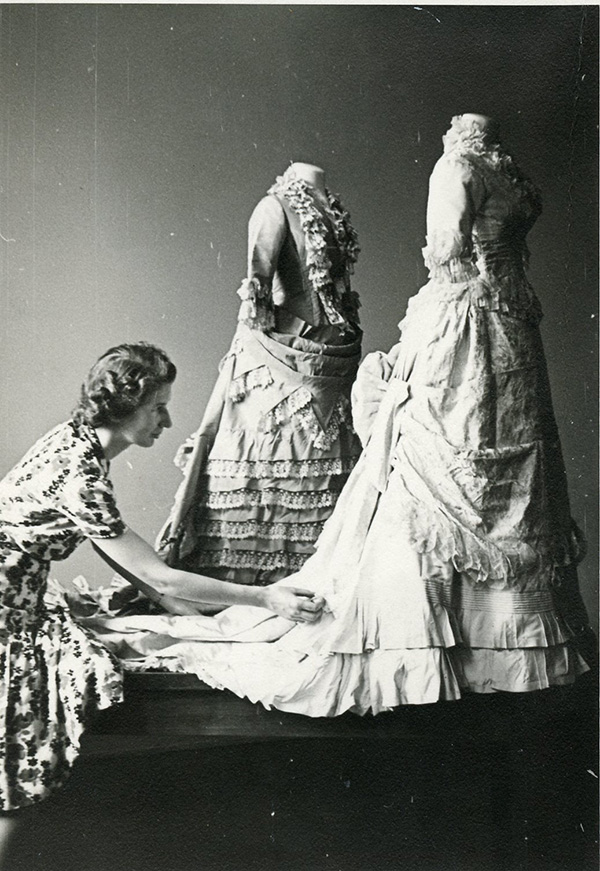 A woman adjusts the train of an ornate, layered gown displayed on a mannequin beside two other mannequins wearing similar vintage dresses.