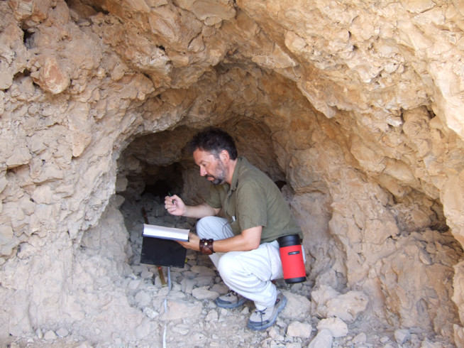 Man wearing glasses crouching inside a rocky cave or tunnel holding a notebook and pen.