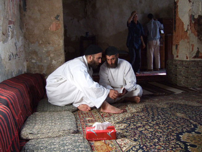 Two men dressed in white robes sitting on a ruged floor, arranging objects inside a small red box.