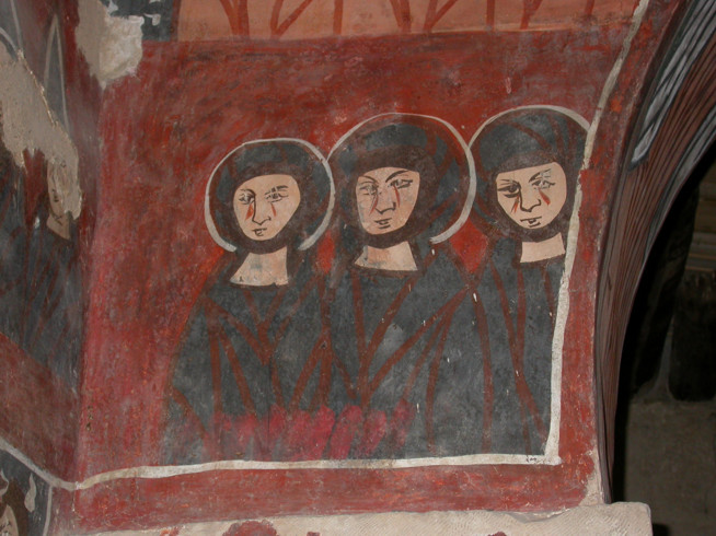 Three medieval figures in dark robes and circular head coverings are painted on a red background on a wall corner.