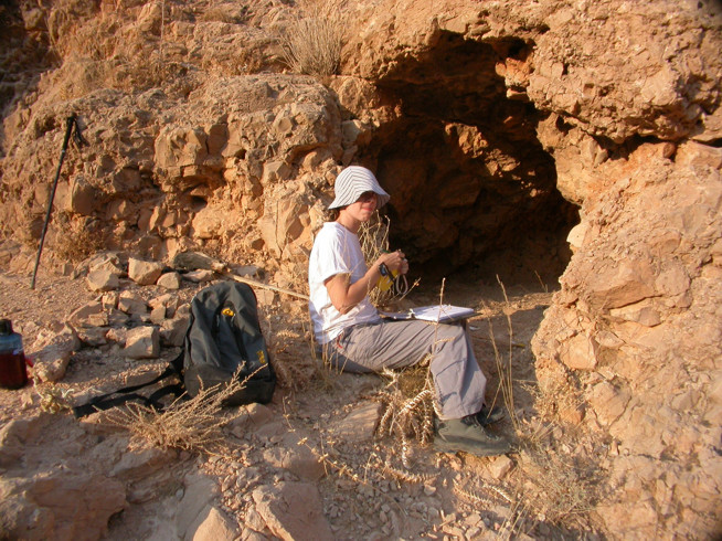A person wearing a white hat and light-colored clothes sits on a rock near the opening of a small cave in a rocky terrain.