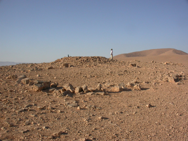 Wide view of ancient stone ruins in a dry, rocky desert landscape with a person standing near the center.