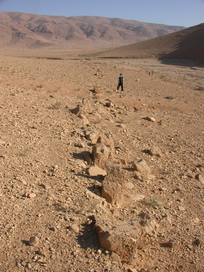 Long, narrow line of large stones arranged in a desert plain with a person standing in the background near distant hills.