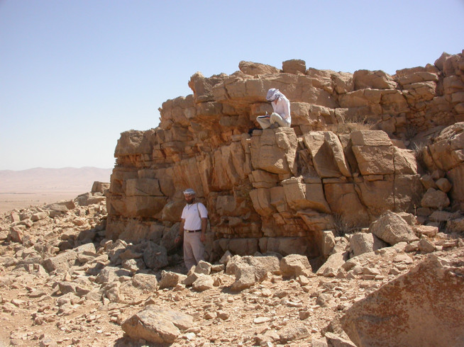 Two people standing and sitting on a large pile of ancient stone blocks in a desert landscape.