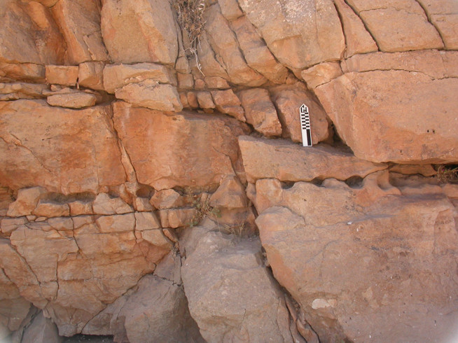 Rock formation with layered, cracked orange-brown stone and a black-and-white striped measuring scale placed on one of the flat surfaces.