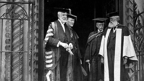 Four men dressed in academic regalia standing on a building entrance decorated with wrought iron gates.